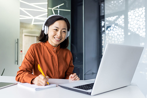 Asian business woman with headphones watching online training course at workplace, woman writing information happy and satisfied with the results of professional development. Photo of a woman at a desk in front of a laptop taking notes while listening to her headphones.